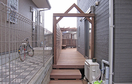 玄関わきのアプローチからのウッドデッキとパーゴラ