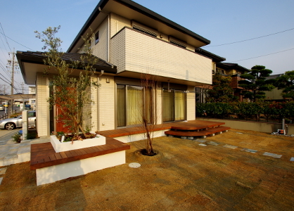 天然木のウッドデッキと繋がった玄関前のベンチ。ベンチで囲まれた花壇にはシンボルツリーのオリーブを配置。<br />
目隠しを兼ねるため、オリーブにしては珍しい株立のような樹形です。